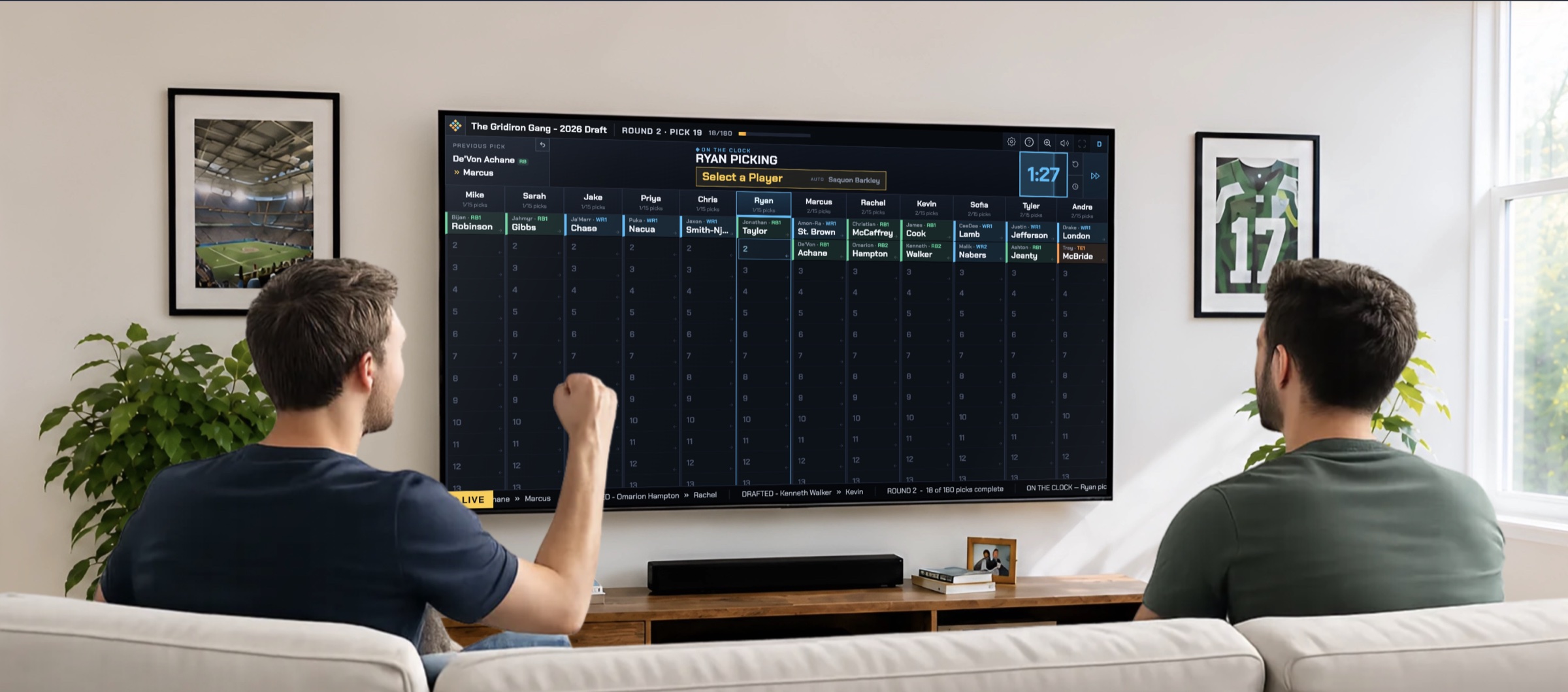 Two friends watching a live FF Drafter salary-cap auction on the TV board in their living room, with a player on the block